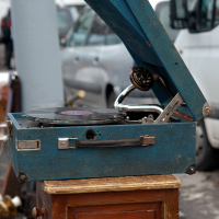 A vintage blue gramophone with a record on the turntable sits on a small brown wooden table. The gramophone's lid is open, revealing the horn and tonearm. In the background, a white van and another vehicle suggest an outdoor setting like Kazjukas 2012 flea market or street sale.