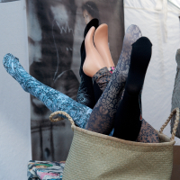 A woven basket tipped over reveals several mannequin legs protruding in different directions. The legs are adorned with various tights and stockings, featuring patterns such as floral, paisley, and solid black. The scene is set against a backdrop with a partially visible image and white fabric, reminiscent of Kazjukas 2012.