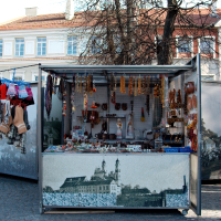 A street-side kiosk displays various amber jewelry, leather crafts, traditional hats, and Kazjukas souvenirs. It has a backdrop depicting a historic cityscape from 2012. There is a stone-paved street in front, and the kiosk is set against a background of buildings and bare trees.