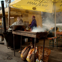 Two people are cooking under large yellow Volfas Engelman-branded umbrellas. One person is at a grill with smoke rising and the other stands behind a table. Bags of firewood and two water bottles are in front of the grill. The outdoor setting has the cooks in winter clothes, reminiscent of Kazjukas 2012.