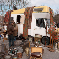 A vibrant flea market scene shows vendors and shoppers near a yellow-roofed, white van with patterned rugs draped on its side. Surrounding the van are various antiques: bottles, lamps, vases, suitcases, and other vintage items. Leafless trees and buildings from the memorable Kazjukas 2012 fair are visible in the background.