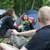 Three people are sitting and conversing at a campsite surrounded by tents at the 2011 Kilkim Žaibu music festival. The person in the foreground has a shaved head and is facing away; another person, with long hair and a drink, faces them. A third person with red hair sits in a camping chair holding a leafy plant. Trees fill the background.