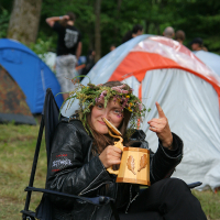 A person wearing a floral crown and a black leather jacket sits in a camping chair at the 2011 Kilkim Žaibu festival. They are holding a wooden mug with a leaf design and giving a thumbs-up. Tents are set up in the background, and other people are visible standing and walking in the wooded area.