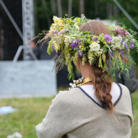 A person with long braided hair stands facing away, adorned with a large, colorful flower crown. They are wearing a traditional light gray garment and a beaded necklace. The background includes a grassy area with trees and some metal stage structures, reminiscent of Kilkim Žaibu 2011, suggesting an outdoor festival setting.
