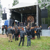 A small crowd of people, many dressed in black leather jackets, stand and watch a rock band performing on an outdoor stage at the 2011 Kilkim Žaibu festival. The stage has large speakers and lighting rigs. Trees and grass surround the area, creating a natural setting. Some people are taking photos.