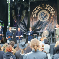 A group of six men dressed in medieval-style clothing stands on a stage surrounded by instruments and audio equipment. They face an audience dressed in casual, modern attire. The backdrop features a large logo and the text "Antiytila" with Viking symbols, echoing the spirit of Kilkim Žaibu 2011. Surrounding trees are partially visible.