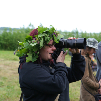 A person with dark hair, wearing a black hoodie and an elaborate green leafy crown, looks through a large camera lens in an outdoor field during the 2011 Kilkim Žaibu festival. Other individuals are blurred in the background, suggesting a group event in a natural setting. The sky appears overcast.