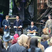 A band dressed in traditional medieval-style clothing performs on a stage at Kilkim Žaibu 2011, using period instruments. The audience, some wearing flower crowns, watches attentively. The background features musical equipment, including Marshall amplifiers and a drum set. The stage is adorned with a large banner.