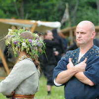Two individuals participate in an outdoor historical reenactment at Kilkim Žaibu 2011. The woman on the left wears a flower crown and a traditional dress, her back to the camera. The man on the right stands with arms crossed, wearing traditional attire. A rustic tent and other participants are visible in the background.