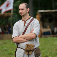 A man dressed in medieval attire stands with folded arms. He wears a light gray linen tunic, a brown leather belt with a pouch, and a shoulder strap holding a wooden object. Behind him is a blurred background of greenery and a flag with a red emblem, reminiscent of Kilkim Žaibu 2011.