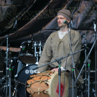 A person wearing traditional beige and brown attire, including a hat, stands while playing a large drum on the Kilkim Žaibu stage in 2011. They are surrounded by microphones with a drum set visible in the background. The person's expression is focused, and the crowd's heads are visible in the foreground.