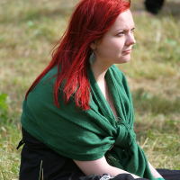A red-haired woman wearing a green shawl over a black dress sits on grass at Kilkim Žaibu 2011. She looks towards the distance with a thoughtful expression. The background includes more grass and the legs of another person, which are partially visible, suggesting an outdoor environment, possibly a park.