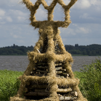 A large, intricately designed bonfire structure from the 2011 Kilkim Žaibu festival stands proudly on a grassy area near a body of water. Made of stacked logs and covered in straw, the conical structure features three prominent straw arches at the top, resembling a crown. Trees and a cloudy sky provide a picturesque backdrop.