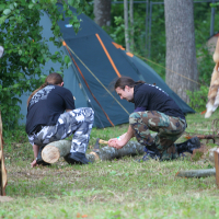 Two people in camo pants and black shirts are kneeling on the ground, chopping a log with an axe in a forested campsite during Kilkim Žaibu 2011. A tent is set up in the background, alongside a pile of fur pelts and wooden structures. The scene is set outdoors with green grass and trees visible.