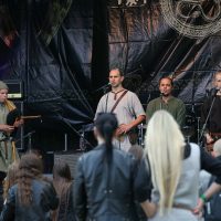 A folk band performs on an outdoor stage at Kilkim Žaibu 2011, featuring four male musicians wearing medieval-inspired attire. One plays a bagpipe, while the others sing into microphones. An engaged audience, some with long hair and dressed in dark clothing, observes the performance. The backdrop is decorated with Celtic patterns.