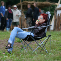 A person with short red hair and glasses relaxes in a black folding chair on a grassy field during Kilkim Žaibu 2011, eating an apple. Wearing a leather jacket, blue ripped jeans, and sandals, they lean back comfortably. In the background, several people are standing and talking near rustic, thatched structures.