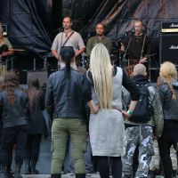 A group of people in various styles of clothing, some with long hair and some with shaved heads, stand in front of a stage at Kilkim Žaibu 2011, watching a live band perform. The musicians on stage are dressed casually and surrounded by musical equipment. The background features a large banner with intricate designs.