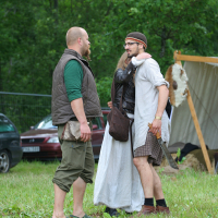 Three people in medieval attire converse outdoors at the 2011 Kilkim Žaibu festival. A bearded man in green and brown stands barefoot. A woman in a white dress and leather vest faces him, arm around a man in a headband, white tunic, and brown pants holding a sheathed sword. A tent and trees are in the background.