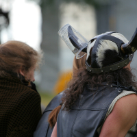 A person dressed in a black leather outfit and a Viking helmet with horns and an attached transparent cup stands facing away at the 2011 Kilkim Žaibu festival. Another person with braided red hair and a brown plaid fabric draped over their shoulders is beside them. The blurred background suggests an outdoor setting.
