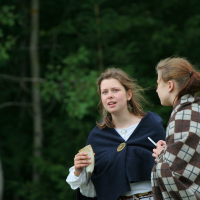Two young women dressed in medieval-style clothing stand in an outdoor setting with lush greenery in the background. One wears a dark blue cloak with a gold brooch and holds a small piece of bread, while the other in a plaid cloak, reminiscent of Kilkim Žaibu 2011, seems to be speaking to her, their expressions engaged.