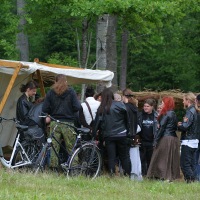 A group of people, mainly dressed in black clothes and leather jackets, gather under a white canopy tent in a wooded area at the 2011 Kilkim Žaibu festival. Two bicycles are parked nearby. There are stacks of hay and men and women engage in relaxed conversation. The overall atmosphere appears casual.