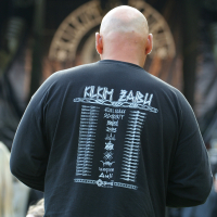 A bald man is seen from behind wearing a black long-sleeve shirt with white text featuring a list of band names and logos in a vertical arrangement. He is at an outdoor setting with a stage in the background, hinting at Kilkim Žaibu 2011. Other attendees are partially visible around him.
