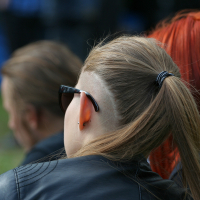 A person with long, blonde hair tied in a ponytail and partially shaved sides is seen from the back. They are wearing sunglasses and a black leather jacket, reminiscent of the Kilkim Žaibu festival in 2011. Next to them is another person with vibrant red hair. Both are seated outdoors, with a blurred background.