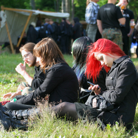 A group of four people dressed in black attire, including one with vibrant red hair, sit on low grass at the 2011 Kilkim Žaibu festival. One person examines a phone while others seem engaged in conversation and activities. In the background, more people are gathered near a fabric-covered shelter in an outdoor setting.