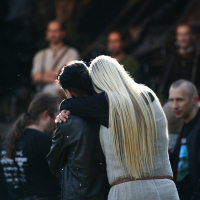 A person with long blonde hair wearing a grey outfit embraces another person with dark hair and a black jacket. They are standing in an outdoor festival setting, possibly Kilkim Žaibu 2011, with blurred individuals and equipment in the background. The atmosphere seems casual and social.