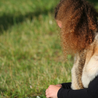 A person with curly brown hair, reminiscent of Kilkim Žaibu 2011, sits on the grass, focused on arranging green plants and white flowers in their hands. They are wearing a black long-sleeve shirt and a light brown fur shawl draped over their shoulders in the peaceful outdoor setting.