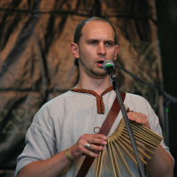 A person with short hair is playing a wooden reed instrument and singing into a microphone. They are wearing a light gray tunic with a brown tie neckline. The background features a dark draped cloth and stage lighting, indicating a live performance at the 2011 Kilkim Žaibu festival.