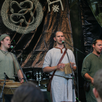 Three men dressed in medieval-style clothing perform on stage at Kilkim Žaibu 2011, each playing a different musical instrument. They are positioned in front of a large tapestry displaying a circular, intertwined snake design. One plays a drum, another holds a small box-shaped instrument, and the third has no visible instrument.
