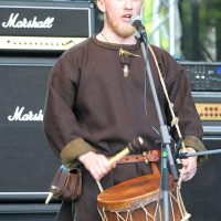 A bearded man is performing on stage at Kilkim Žaibu, 2011, holding a drum and a drumstick while singing into a microphone. He is dressed in historical or medieval-style clothing, featuring a brown tunic and red trousers. Behind him are two Marshall amplifiers. The background includes stage equipment and a metal structure.