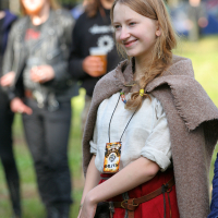 A young woman with blonde hair, wearing a beige shawl over a white shirt and red skirt, smiles outdoors at the 2011 Kilkim Žaibu festival. She has a pouch and a festival pass around her neck. Several people in the background, dressed casually, are out of focus. Trees and greenery are visible, indicating a nature setting.