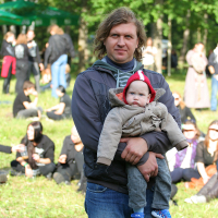 A man with shoulder-length hair in a gray hoodie holds a young child wearing a red hat and beige coat. They are standing on grass in a park during the 2011 Kilkim Žaibu festival, with trees in the background. Several people dressed in black are seated on the grass, engaging in conversations and activities.