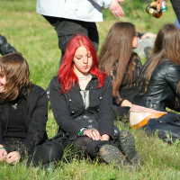 A group of people sitting on a grassy field at the 2011 Kilkim Žaibu festival. In the foreground, two people in casual clothes sit on the grass; one with black and red hair, and the other with brown hair. Other individuals are scattered in the background, some sitting, some standing, wearing dark-colored clothing.