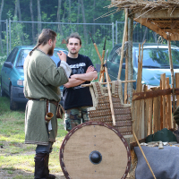Two men are standing beside a medieval-style market stall with various wooden items at the Kilkim Žaibu festival in 2011. One man, in historical attire with a green tunic, brown belt, and boots, holds a cup. The other, in a black t-shirt and camouflage pants, stands with arms crossed. A round shield rests in front of the stall. Trees and parked cars