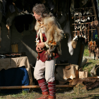 A man dressed in historical attire stands at an outdoor market. He wears a brown tunic, tan pants, and tall brown leather boots with red laces. Various animal pelts drape over his shoulders, and behind him, there are numerous animal pelts and handcrafted items displayed in a wooden stall at the 2011 Kilkim Žaibu festival.