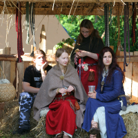 Four people in medieval costumes sit and stand in a rustic, open wooden shelter with a thatched roof at the 2011 Kilkim Žaibu festival. Two women in red and blue dresses sit on straw bales, one holding a drink. Another woman in red stands beside them while a man in camouflage pants sits nearby. Colorful ribbons hang in the background.