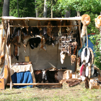 A market stall with a wooden frame and canvas roof displays various fur pelts, hides, and leather goods. The items, in different sizes and colors, hang from the frame and are spread on tables and boxes below. Set in a wooded area with green trees and shrubs in the background, it feels like a scene from Kilkim Žaibu 2011.