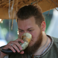 A bearded man dressed in traditional historical attire drinks from a horn-shaped vessel under a thatched roof structure at the 2011 Kilkim Žaibu event. He appears focused, holding the vessel close to his mouth. The background is slightly blurred, indicating an outdoor setting with natural light.