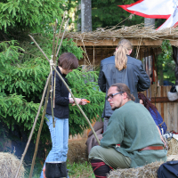 A group of individuals in medieval-style attire are gathered in a rustic outdoor setting, evoking the spirit of a 2011 Kilkim Žaibu festival. One person tends to a small campfire with a pot hanging above it, surrounded by hay bales. Behind them is a simple wooden shelter with a thatched roof. Trees and greenery fill the background.
