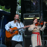 A man and a woman perform on an outdoor stage at Kilkim Žaibu 2011. Both are dressed in medieval-style clothing. The man plays an acoustic guitar and sings into a microphone, while the woman plays the violin. Behind them is a large Marshall amplifier and a black backdrop, with trees visible in the background.
