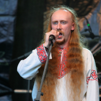 A man with long, red hair and a beard sings into a microphone. He is wearing a traditional, embroidered white shirt with red and black patterns around the collar and sleeves, reminiscent of styles seen at Kilkim Žaibu 2011. A matching embroidered belt completes his outfit. The background is dimly lit, focusing attention on the singer.