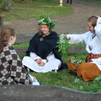 Three women in traditional clothing sit on the grass inside a wooden fence at the 2011 Kilkim Žaibu festival. One, wearing a flower crown, smiles with closed eyes. The second adjusts her hair, and the third has her back to the camera, wrapped in a plaid blanket. Leaves and branches lie scattered around them.