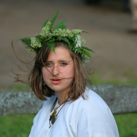 A young woman with light brown hair wears a white garment and a wreath made of green leaves and small white flowers. Seated on a wooden bench, she looks slightly to the side. Her attire and accessories suggest she may be participating in a cultural or traditional event like Kilkim Žaibu 2011.
