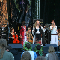 At the 2011 Kilkim Žaibu festival, a band performs on an outdoor stage, featuring members playing cello, guitars, and traditional instruments. Four vocalists, wearing various costumes including dresses and long coats, stand at the front of the stage. In the foreground, some audience members, one with a flower crown, watch the performance.