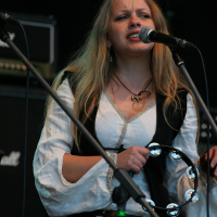 A woman with long blonde hair is performing at the Kilkim Žaibu festival in 2011, holding a tambourine in one hand and singing into a microphone. She is wearing a white peasant-style blouse under a black vest. Behind her, there's audio equipment, including a Marshall amplifier. The stage is dimly lit.