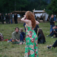 A woman with long red hair, wearing a green floral dress, stands on a grassy field at what appears to be Kilkim Žaibu 2011. She holds her hands near her face while several others sit and stand in the background, with tents and trees indicating an outdoor festival. The atmosphere is relaxed and casual.