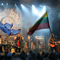 A band performs on stage at an outdoor Kilkim Žaibu 2011 concert, with one musician playing an acoustic guitar. The stage backdrop features the band’s logo. Two large flags, one with Lithuanian colors and another with a different design, wave in the foreground. The audience watches the performance under stage lights.